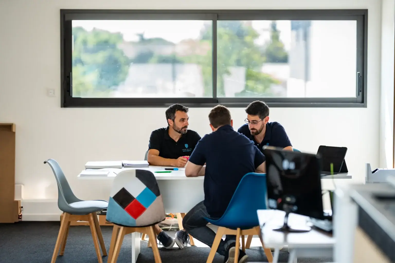 Trois membres de l'équipe Vestalia en réunion autour d'une table, discutant de solutions technologiques pour la maison intelligente, avec des documents et un ordinateur portable visibles.