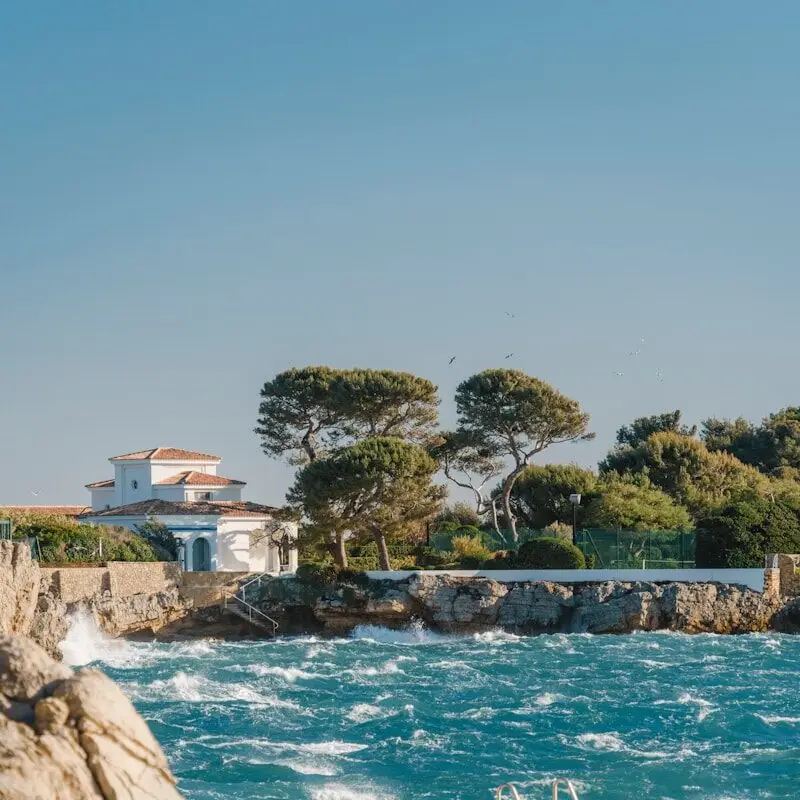 Vue côtière en Provence-Alpes-Côte d’Azur, maison blanche avec toit en tuiles, arbres verdoyants, vagues de mer turquoise, ciel dégagé.